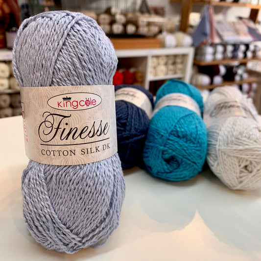 Ball of King Cole Finesse Cotton Silk DK yarn on a table with other yarn balls in the background. Buy now from The Knit Lounge UK yarn shop.